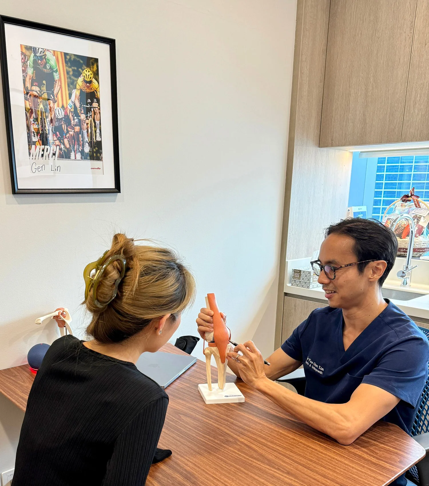 Dr Foo Gen Lin consulting a patient at Apex Sports Clinic Singapore, explaining knee anatomy using a 3D model to diagnose and discuss treatment options for knee pain.